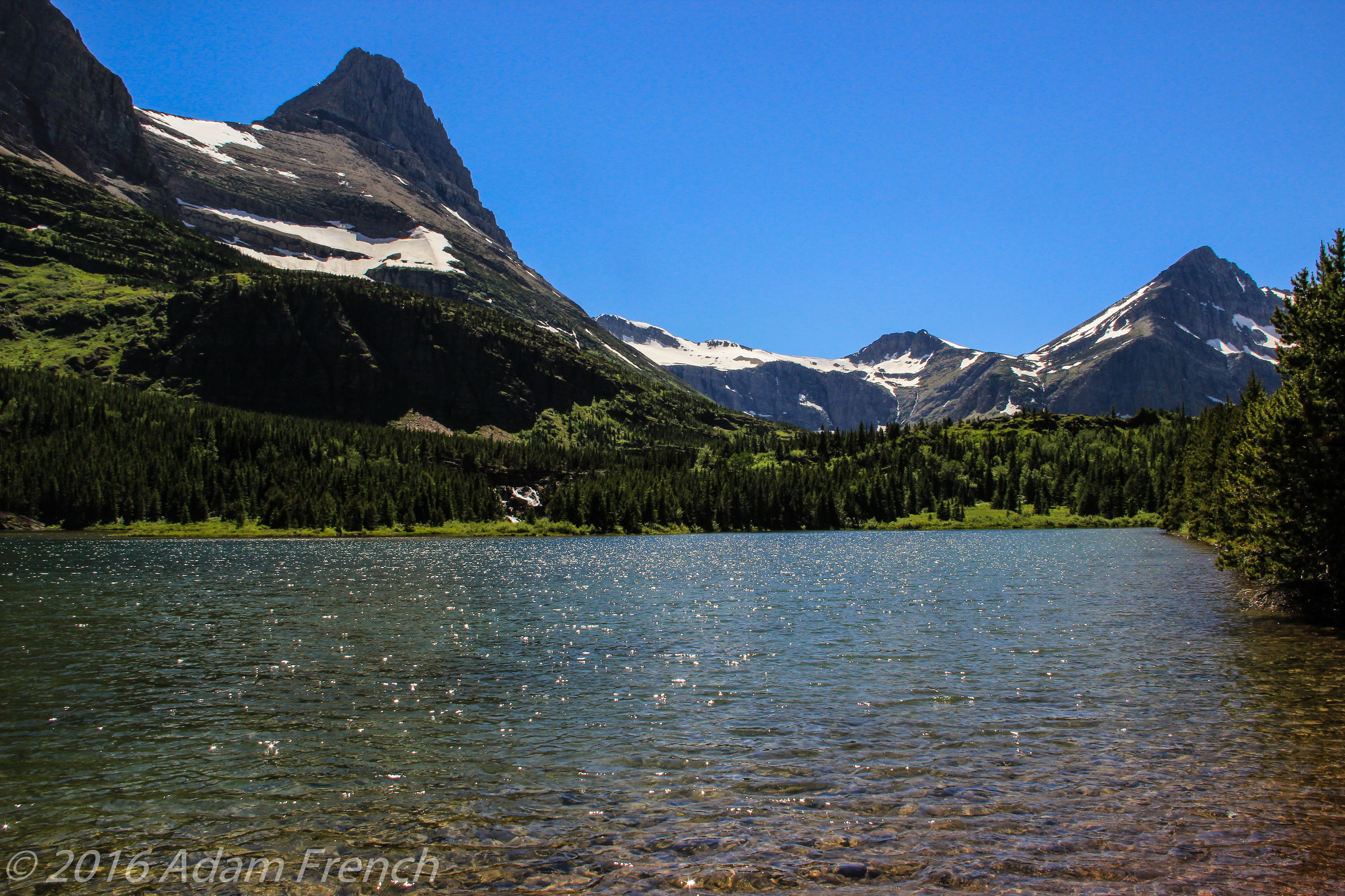 Lakewater ripples surrounded by rocky, towering mountains spotted with snow, all under a clear, blue sky