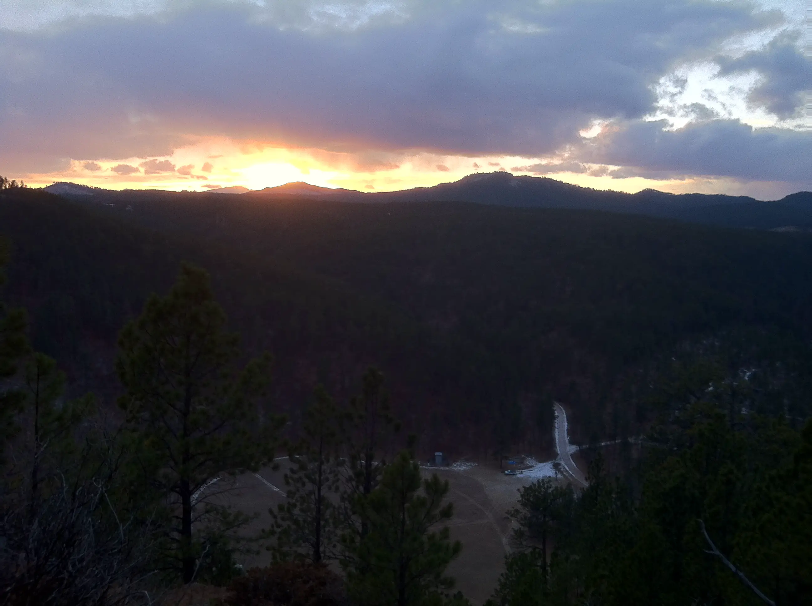 Sunset shot from above over a valley floor. There is a meadow and pine trees at the bottom. The dark silhouette of mountains is in the distance with the sun setting behind them.