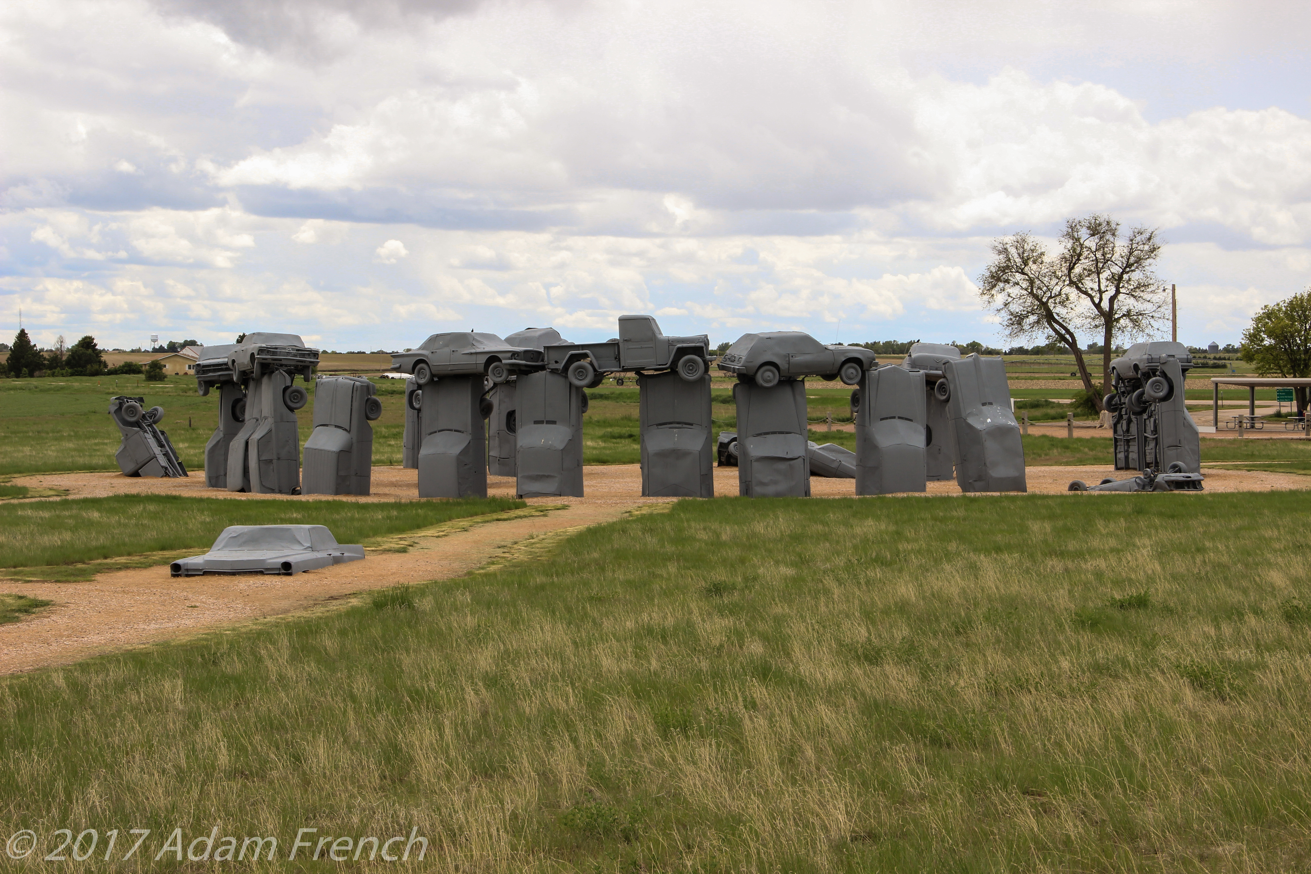 A circle-monument made of gray cars sits in a green field on a dirt path