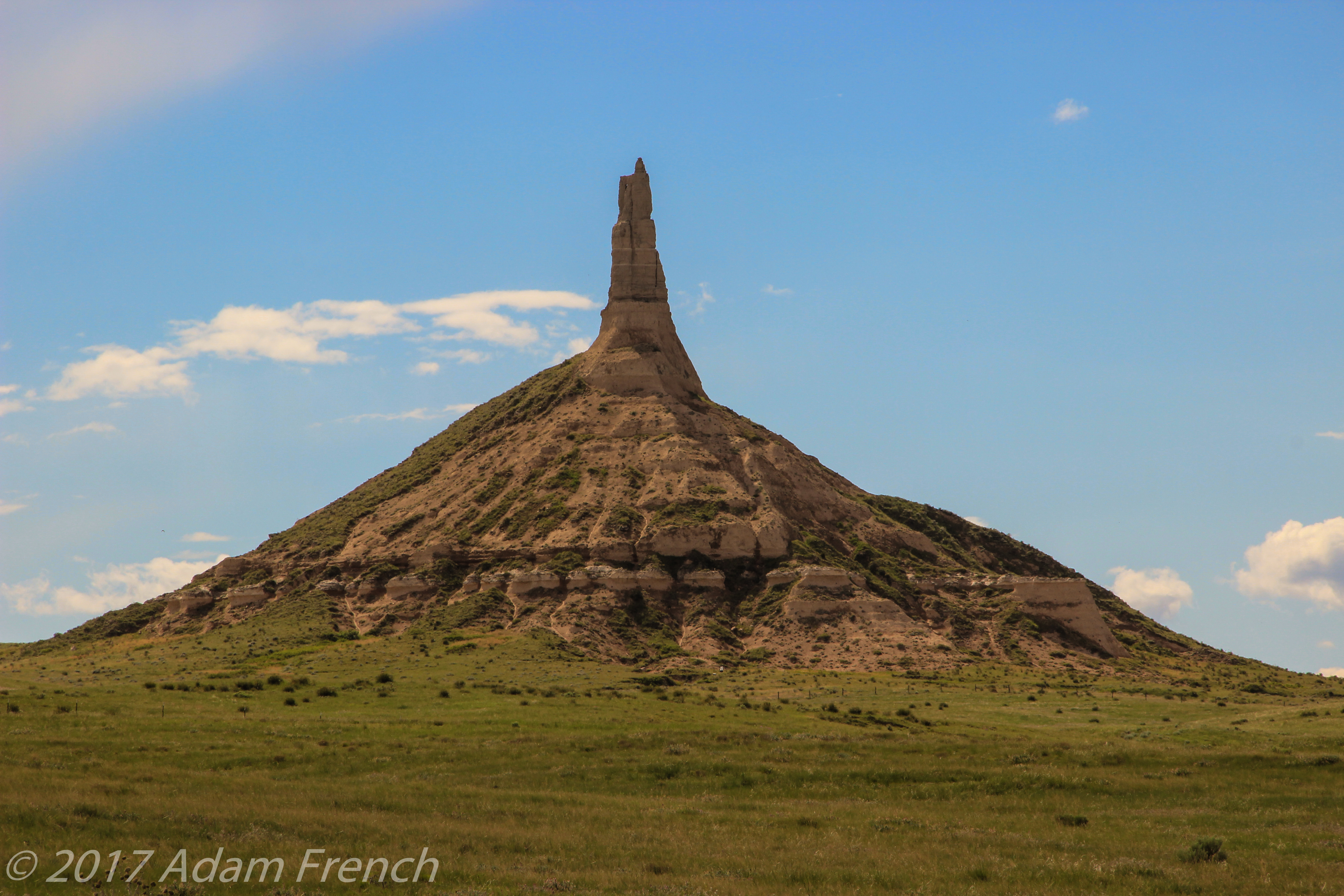 A red rock spire rises from a red rock hill on a green plain