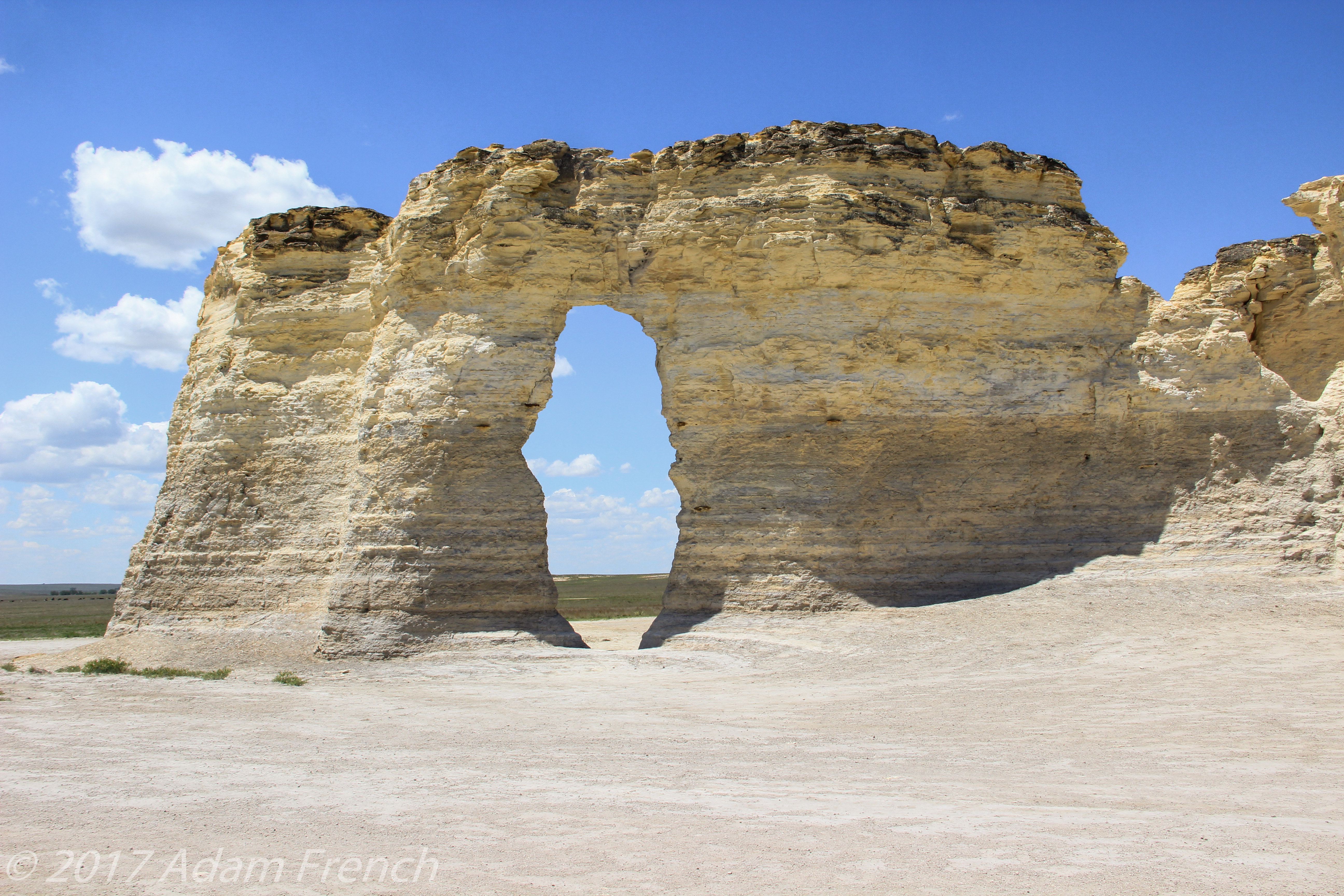 A large, white, rock rises from the ground with a large hole in the middle in a sandy, desert-like area