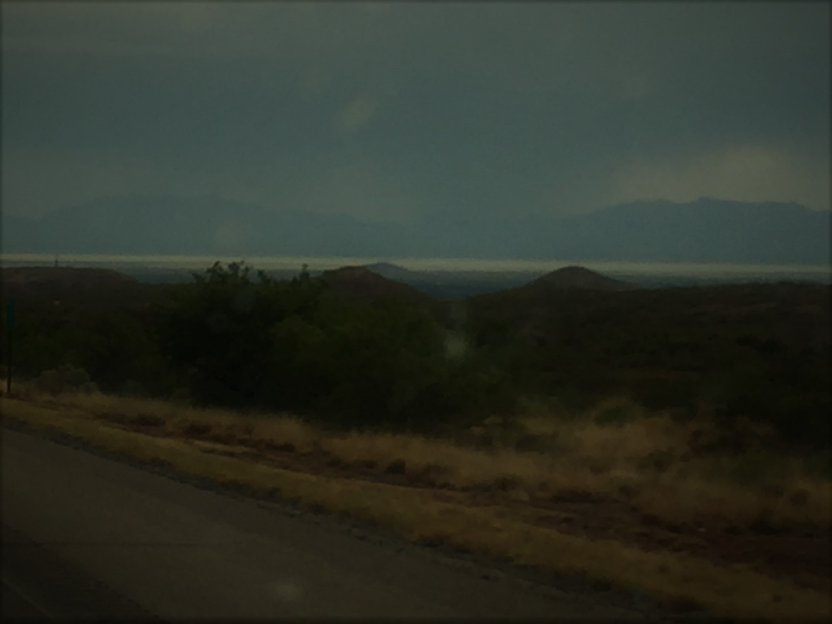 A dark picture where desert scrubland in the foreground and dark mountains in the background are barely visible. In the far background appears to be a white, shimmering lake. 