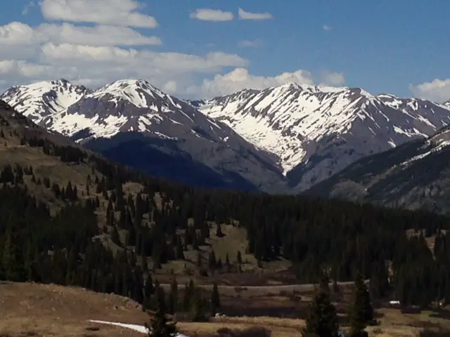 Tree-covered mountain sides in the foreground, snow covered, rocky mountains in the background. 