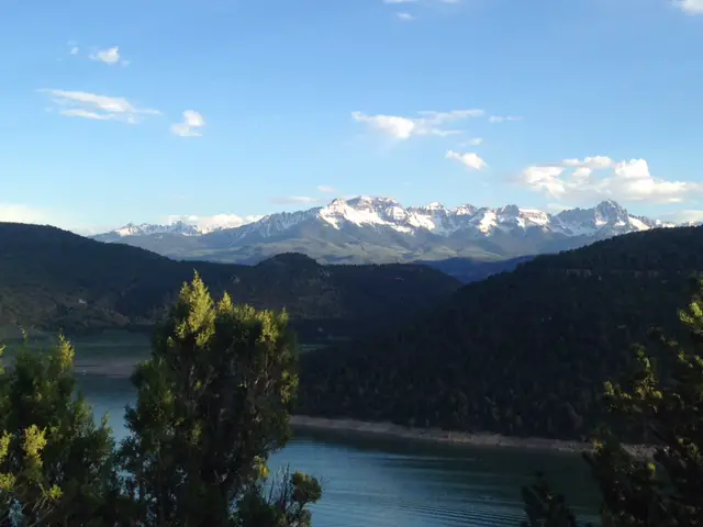 Green trees and a lake in the foreground, tree-covered mountains behind with snow-topped, rocky mountains in the background