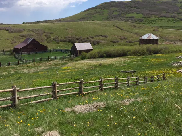Several ranch buildings (barns, an old house) sit in a field behind fences. A grassy hill rises in the background.