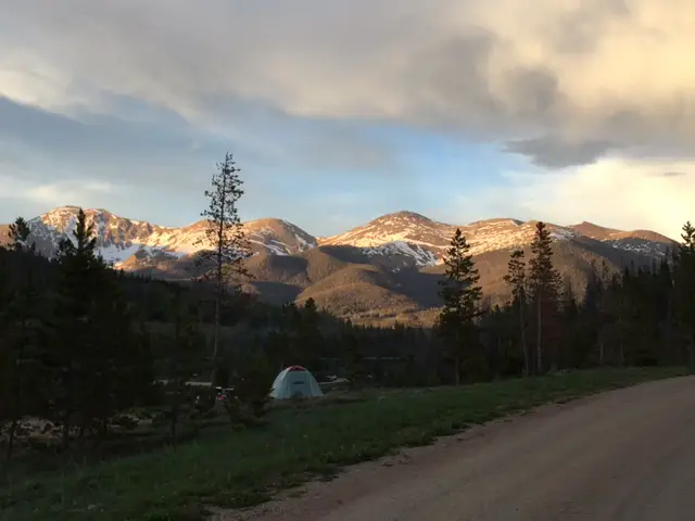 A tent on grass, surrounded by trees in the foreground. Rocky, snow-topped mountains glowing a light pink in the sunset in the background