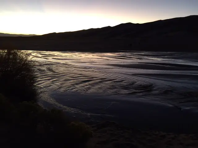 Sunset over a wide, very shallow creek that runs over a sandbar. Dark silhouettes of sand dunes are in the background. 