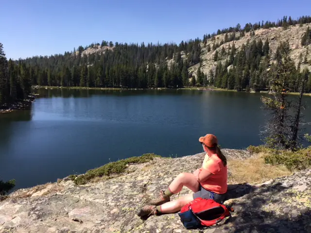 A woman sits looking out over the dark, smooth water of Mirror Lake surrounded by forest. A tree-covered mountainside is in the background.