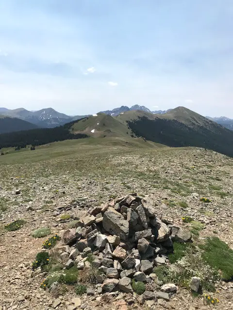 A small, rock cairn in the foreground with a grassy meadow spreading out behind. A spine of grass and tree-covered mountain peaks can be seen in the distance with occasional spats of snow