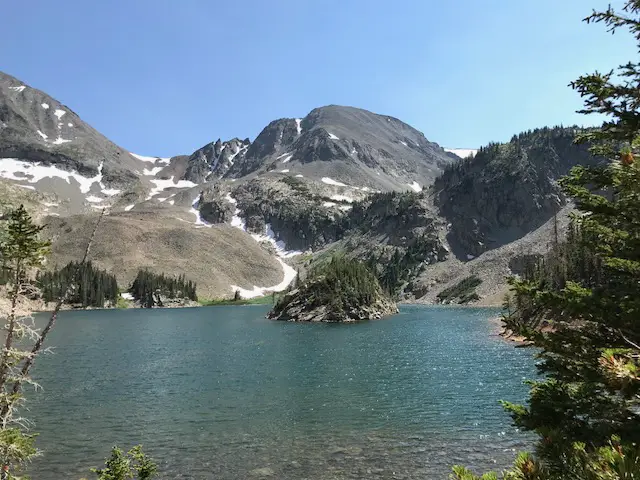 Serene, blue lake surrounded by green, pine trees and rocky mountains with a little snow left on them. All under a clear, blue sky