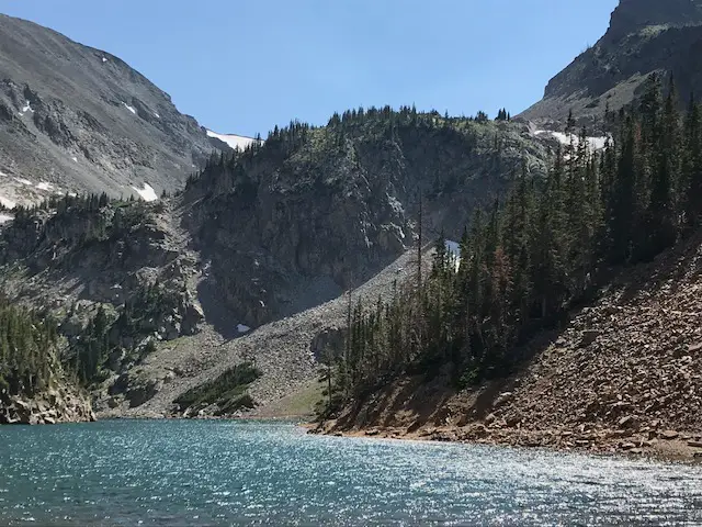 Serene, blue lake surrounded by green, pine trees and rocky mountains with a little snow left on them. Some slopes are covered in scree. All under a clear, blue sky