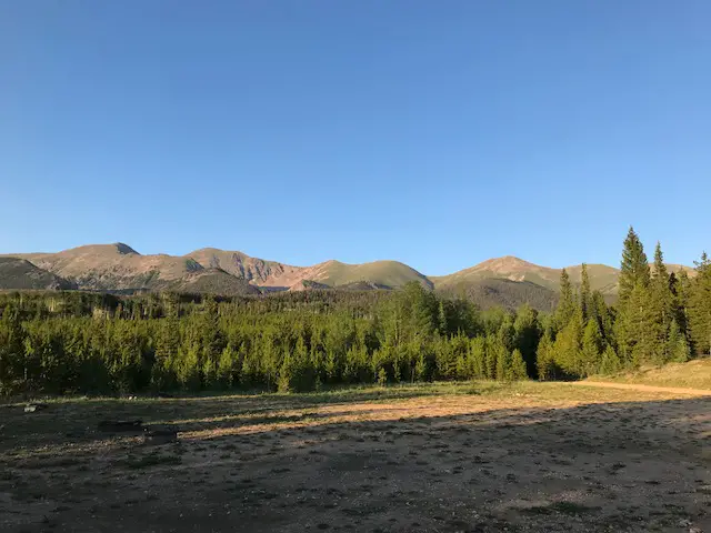 Grassy meadow surrounded by pine trees. Rocky mountains in the background, all under a clear, blue sky.