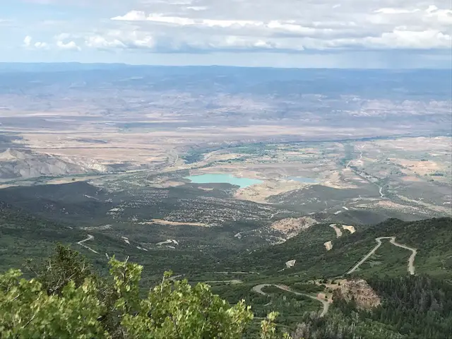 Green tree and grass-covered mountains. Drier scrubland covers the lower elevations in the background. A dirt road traverses the forestland as it descends the mountain. The blue waters of a lake sit in the middle of the drier, lower altitudes in the background 