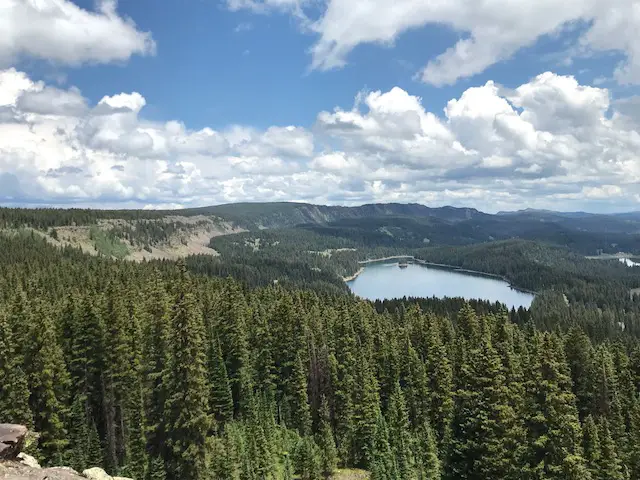 Pine tree-covered mountainside. In the background green trees surround a blue lake with a small, green island in the middle. A craggy, mountain wall is on one side