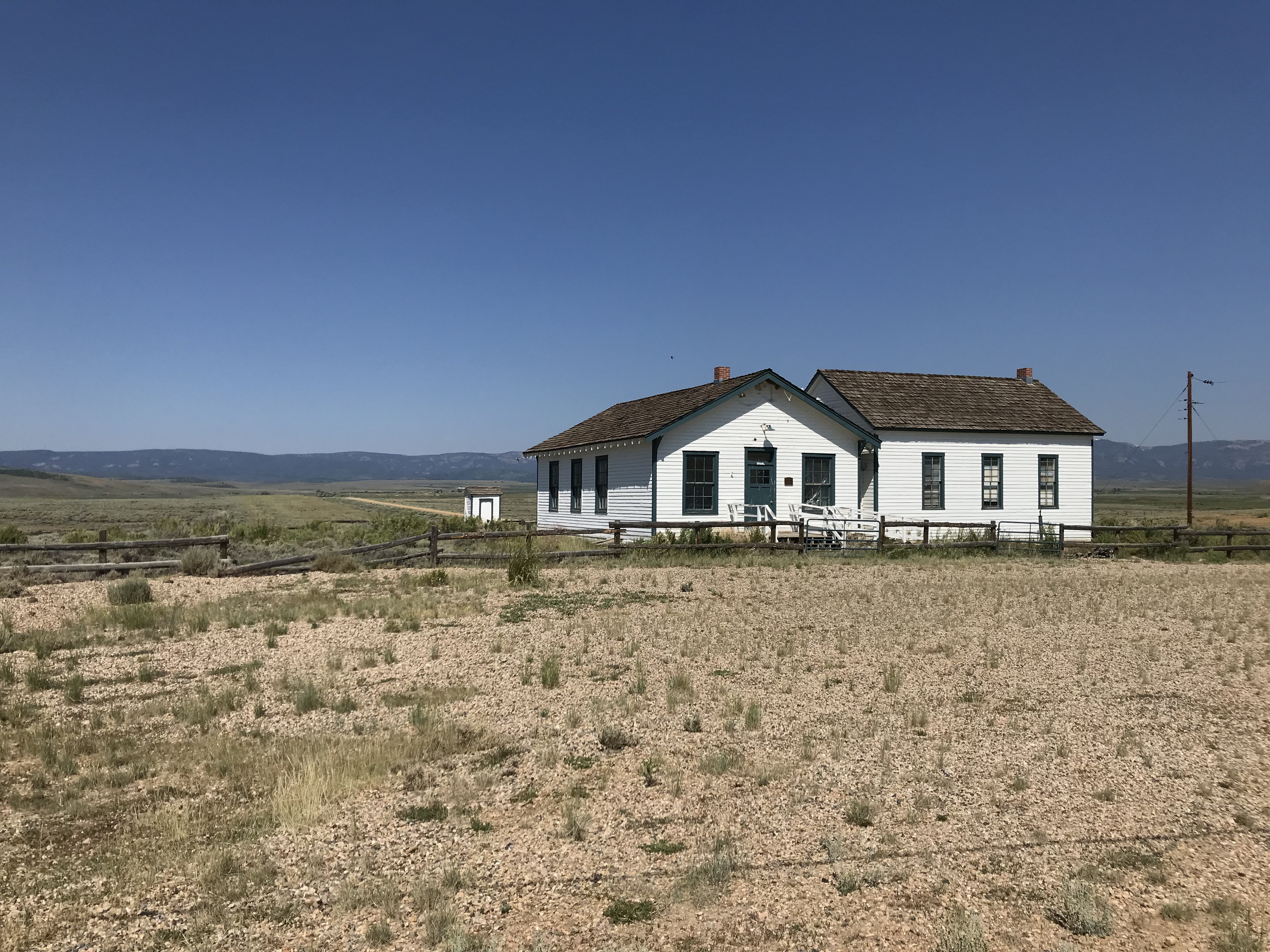 Old, white-boarded schoolhouse building sits in an arid landscape. Dark mountains are seen far in the background