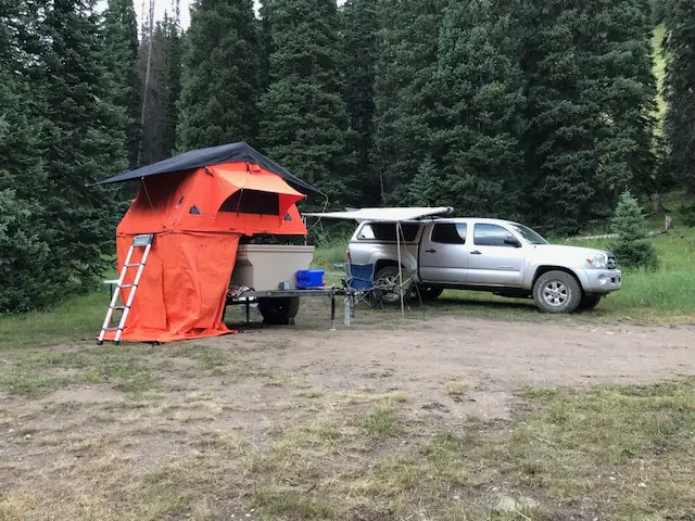 An orange rooftop tent sits on a trailer in front of a truck with an awning. This is in a clearing with tall pine trees surrounding it
