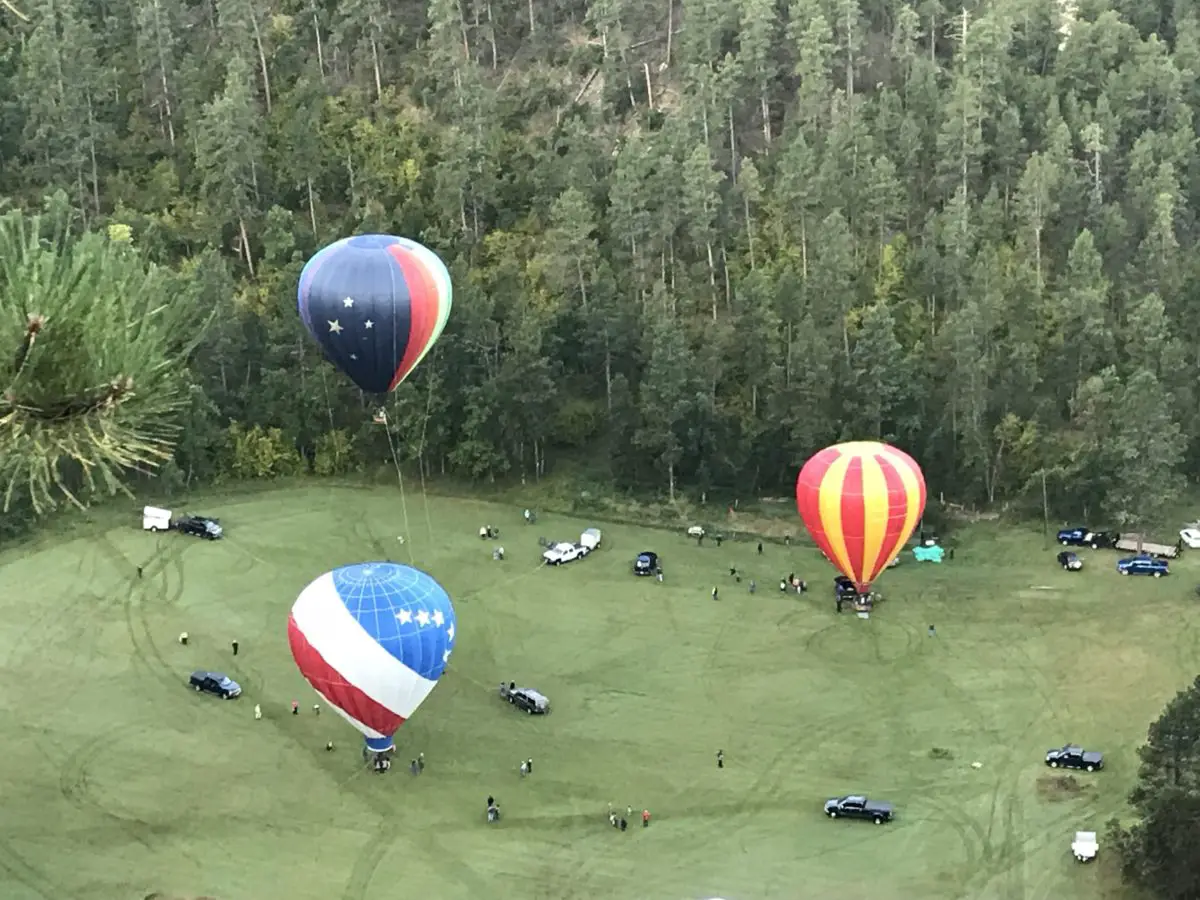 Top view of four colorful, hot air balloons floating near a grassy, valley floor surrounded by pine trees. Cars and trucks also dot the valley floor.