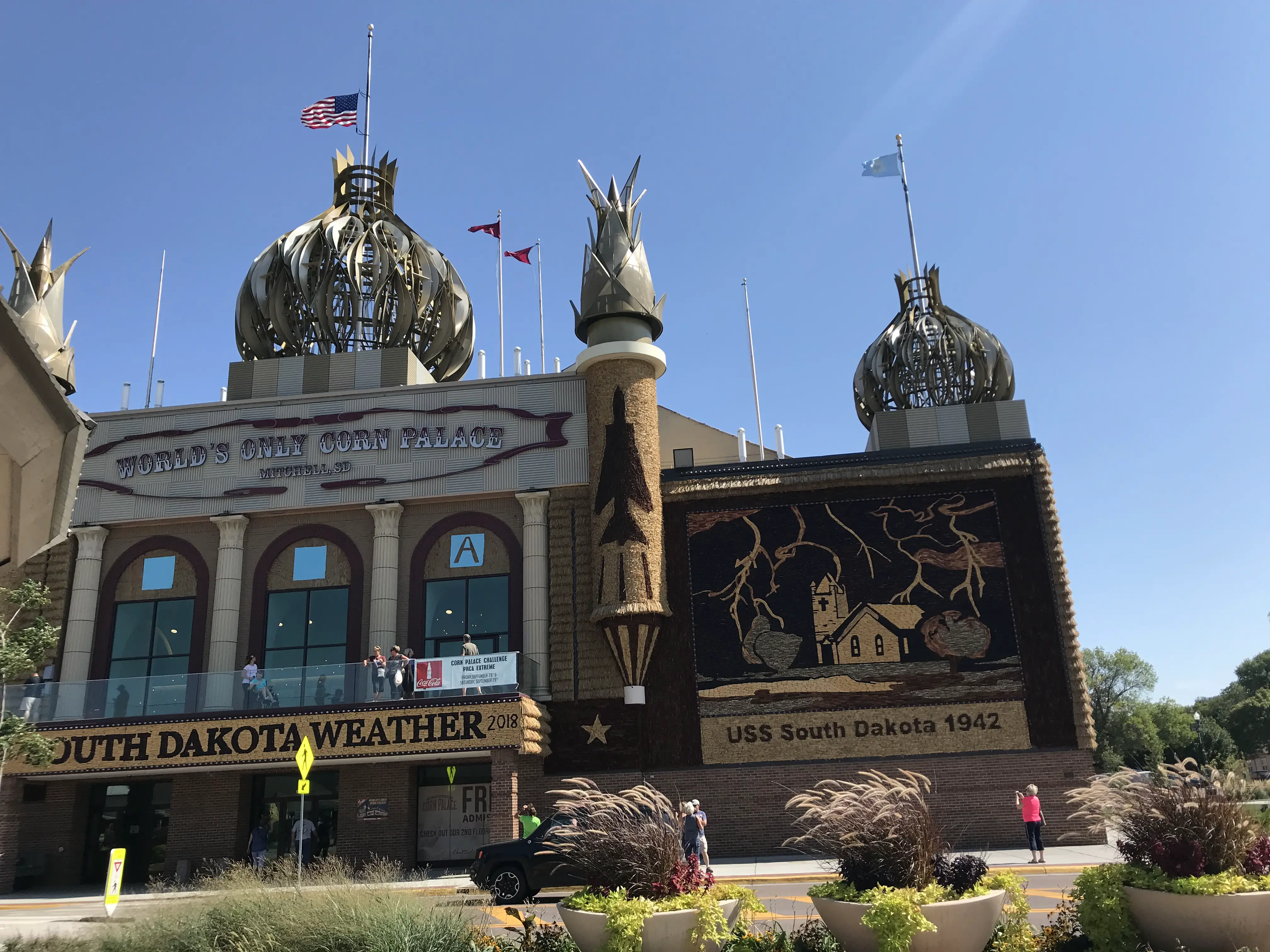 A building that looks like a palace with corn cobs coming off the top of the towers and signs that say, "South Dakota Weather", "USS South Dakota 1942" and "World's Only Corn Palace.