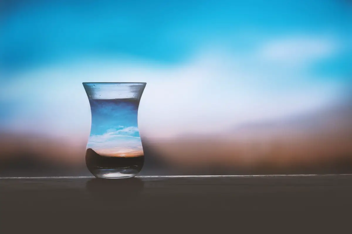 A vase full of water sits on a table and is set against a blending of blue, white and brown colors in the background. The colors are reflected in the water.