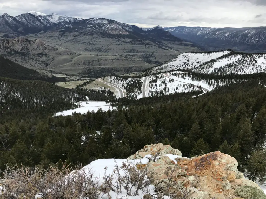 View over a mountain vista from above. Pine trees and snow cover the lower elevations, brown grass covers the higher elevations that leads to tree and snow covered mountains in the distance. 