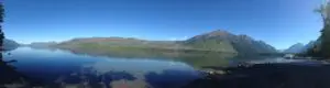 Panoramic view of a flat-water lake reflecting the tree-covered and rocky mountains surrounding it, all under a clear, blue sky.