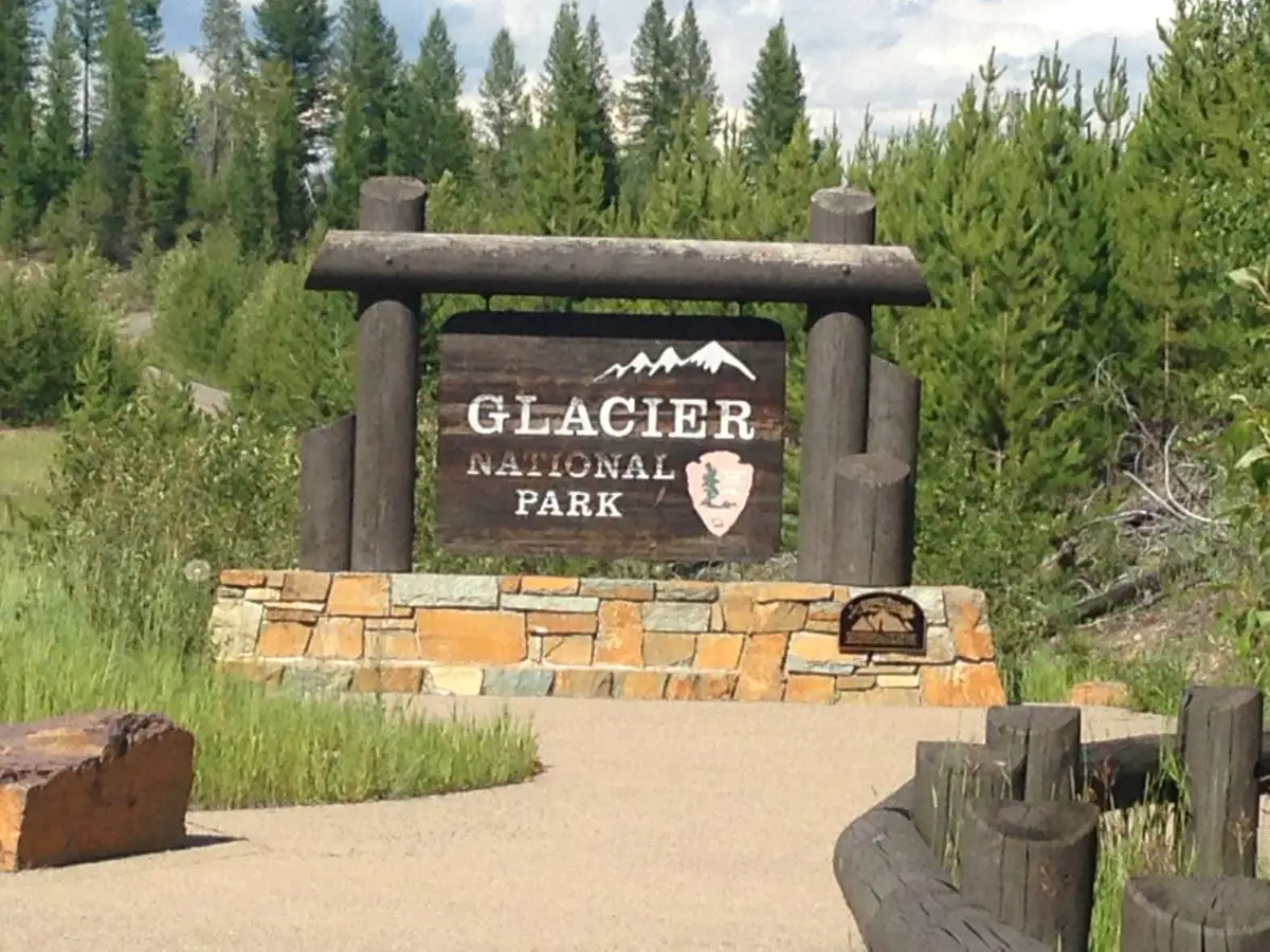 Wooden, national park signs stands on a rock podium in the woods. Sign reads, "Glacier National Park"