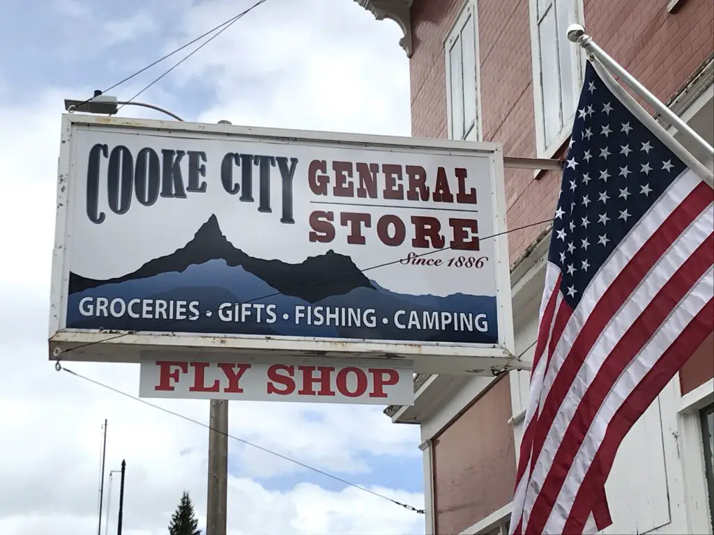 Store sign on a building near an American flag reads, "Cook City General Store, Groceries, Gifts, Fishing, Camping, Fly Shop"