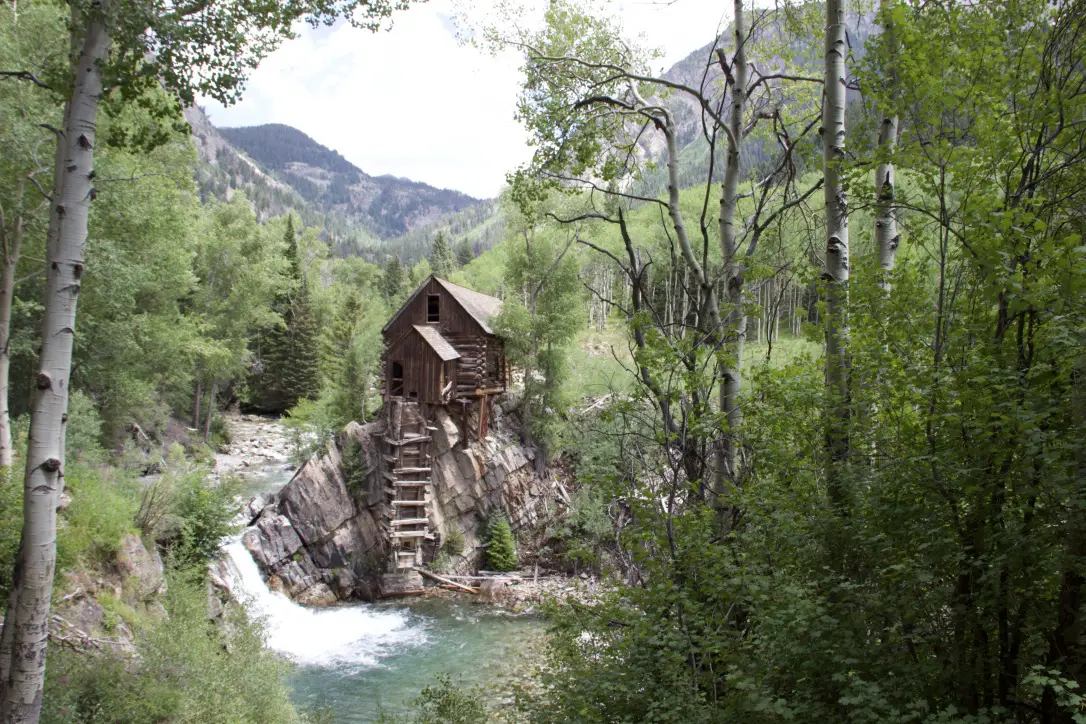 An old, wooden structure sits on a cliff on the edge of a creek. Green trees and a creek surround it. Rugged mountains can be seen in the background