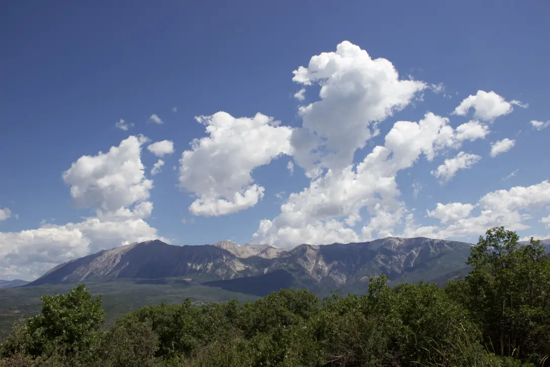 Green trees with rocky mountains in the background and a blue sky with puffy, white clouds