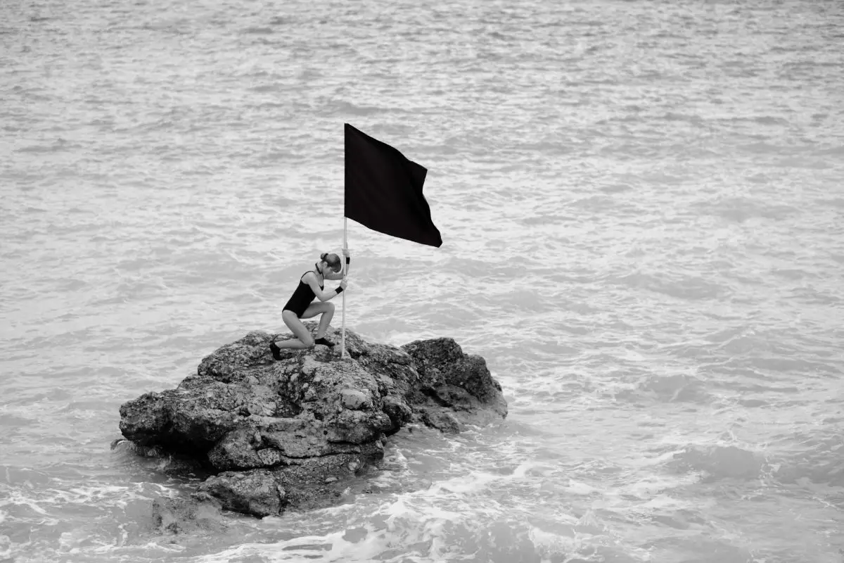 Black and white picture: a person plants a flag on a large boulder surrounded by water