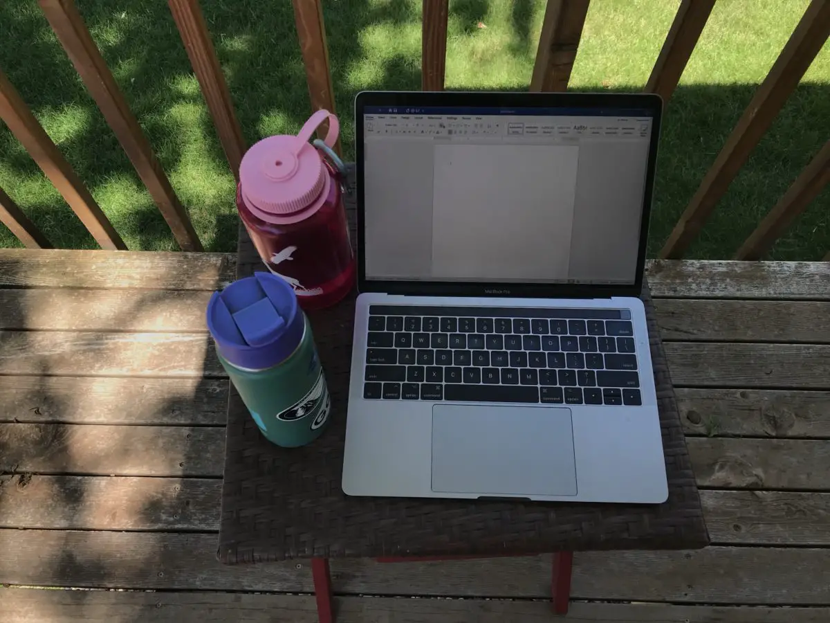 A laptop, water bottle and drink container sit on a table on the deck of a house