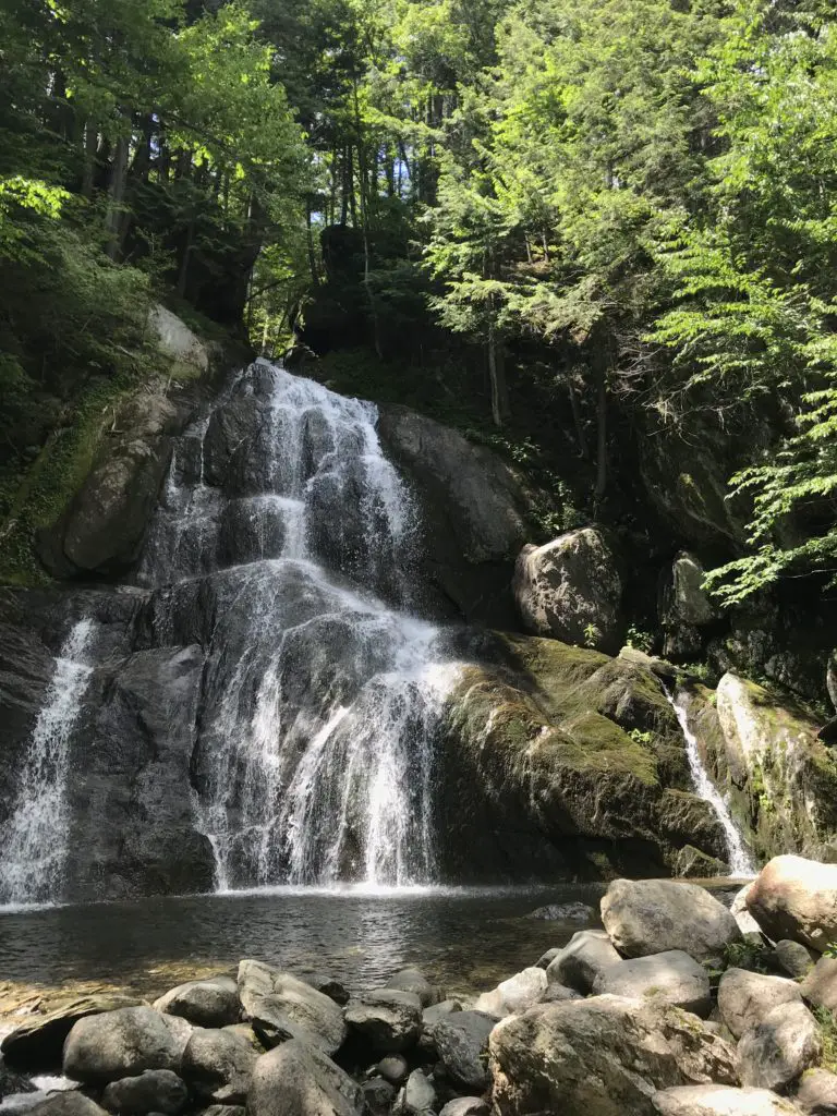 A waterfall over rocks landing in a pool at the bottom in the middle of the woods. Large rocks sit in a pile near the pool of water