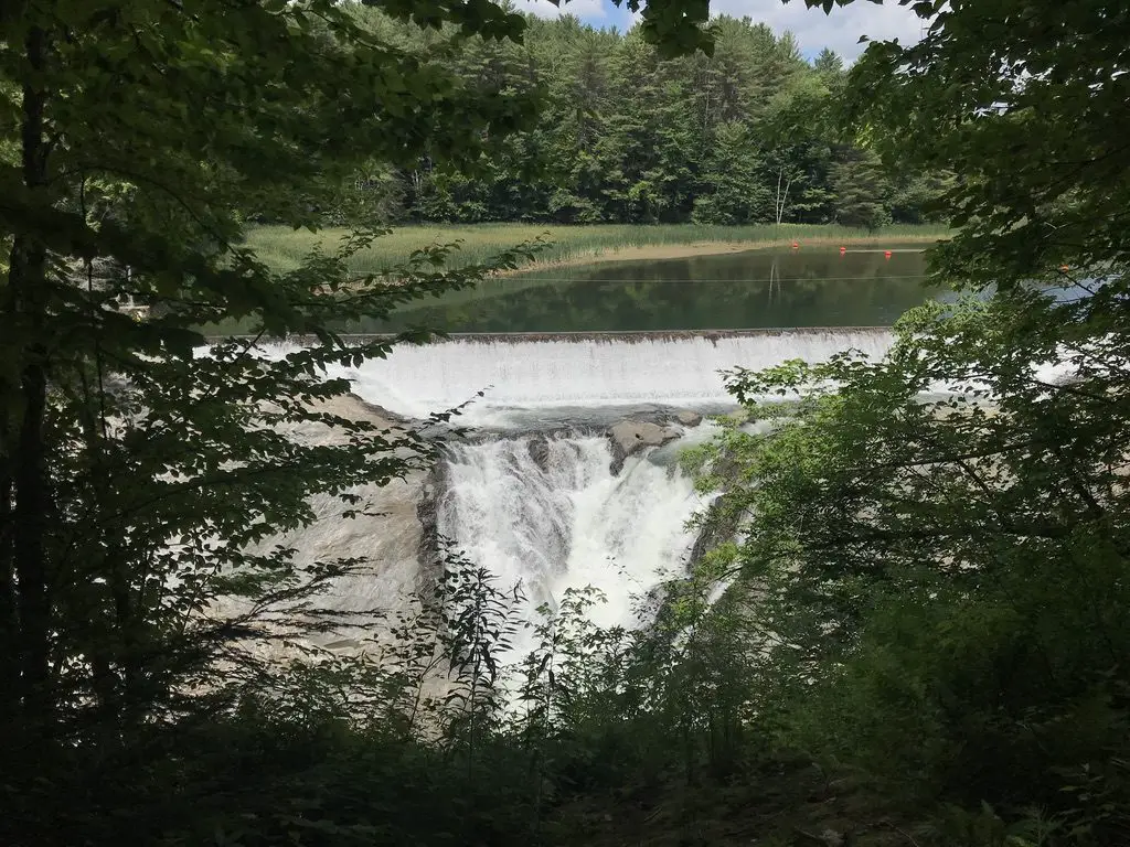 Looking through a hole in the trees, a dam in the background narrows into a large waterfall over some rocks