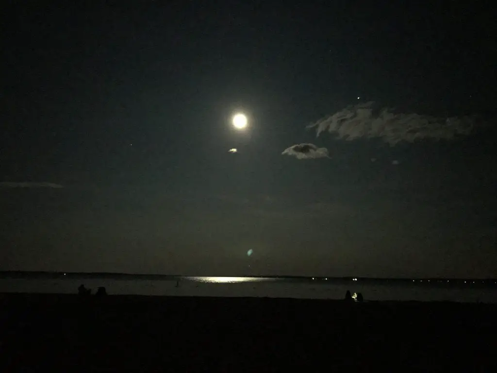 A full moon in the dark, night sky. It is reflected in the dark water of the lake, below