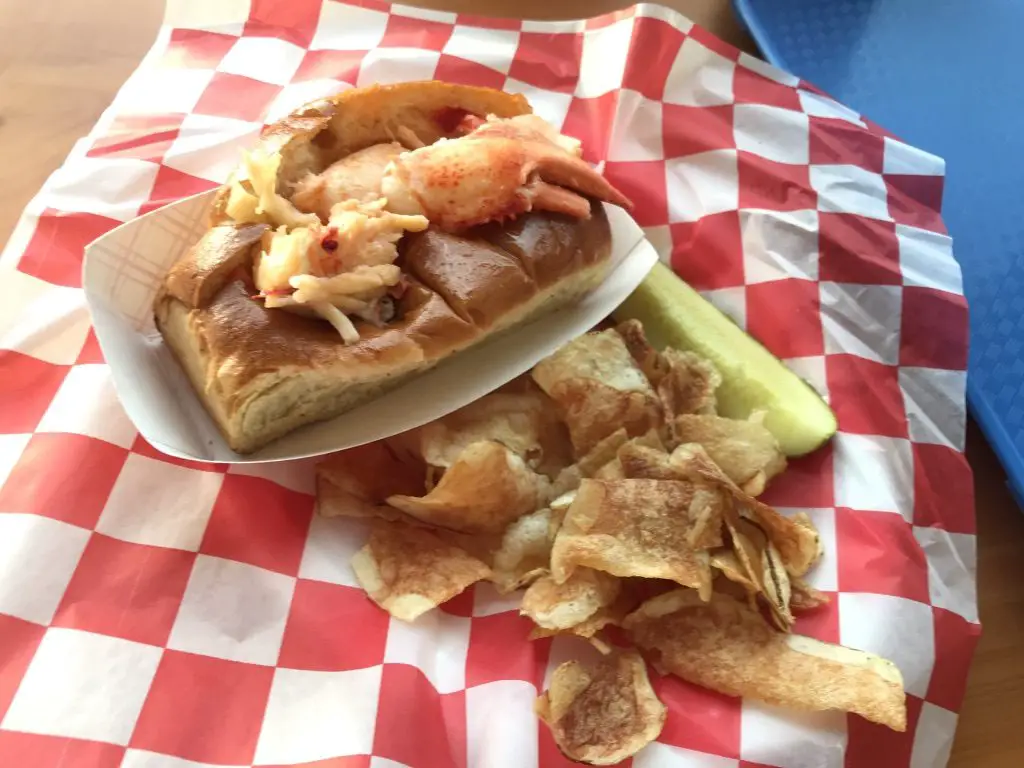 A lobster roll with chips and a pickle on a tray