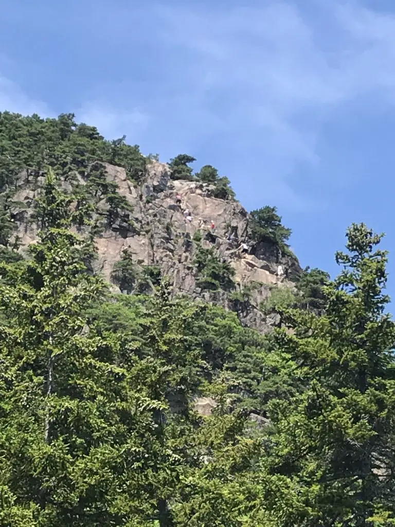 Green trees in the foreground with a rock wall, scattered with trees in the background, all under a blue sky. A line of tiny, multi-colored hikers is climbing the rock wall