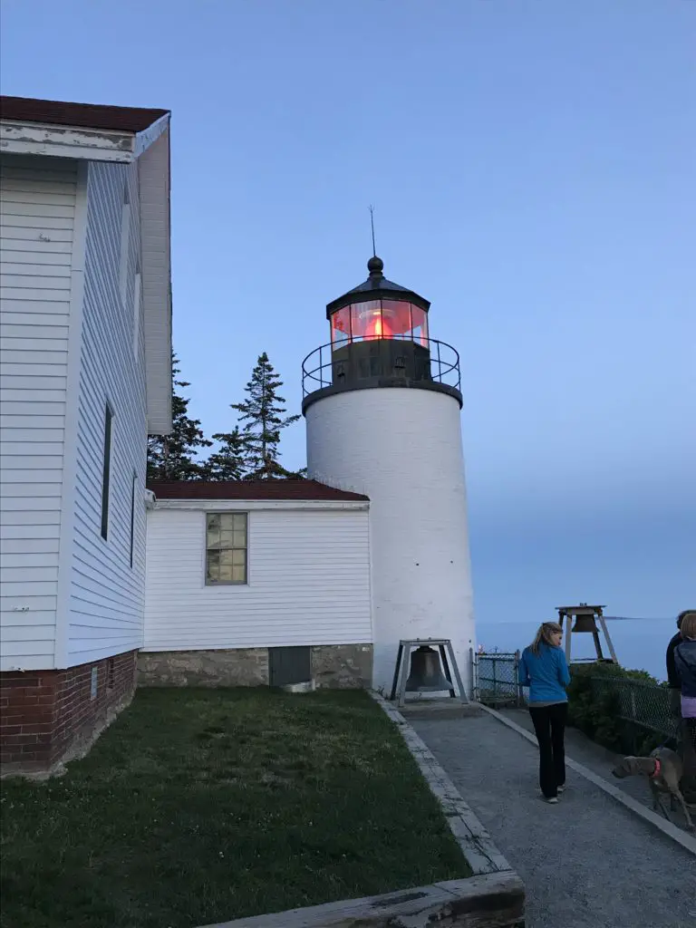 In the darkening light, a white building and lighthouse with a reddish-orange light on top. A bell sits in front of the lighthouse tower