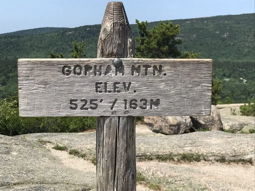 Wooden sign post on a rocky, mountain summit. Green, tree-covered mountains rise in the background. Sign post reads, "Gorham Mtn. Elev. 525'/163 m