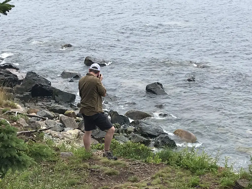 Rear view of a man standing on the rocky shore, taking a picture while looking out into the water