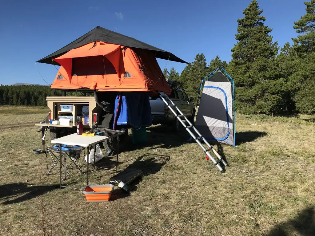 A rooftop tent is open on top of a trailer, a separate small tent and truck are behind. All in a woodland scene.