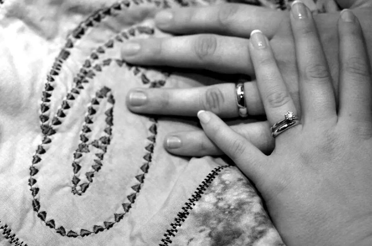 Black & white photo of two hands on a quilt, both wearing wedding rings on the ring fingers