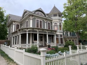 A grey, Victorian-style house sits behind a white, picket fence in town