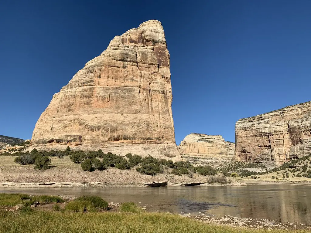 A river with a large rock that resembles a shark's fin rising behind it, all in a desert landscape.