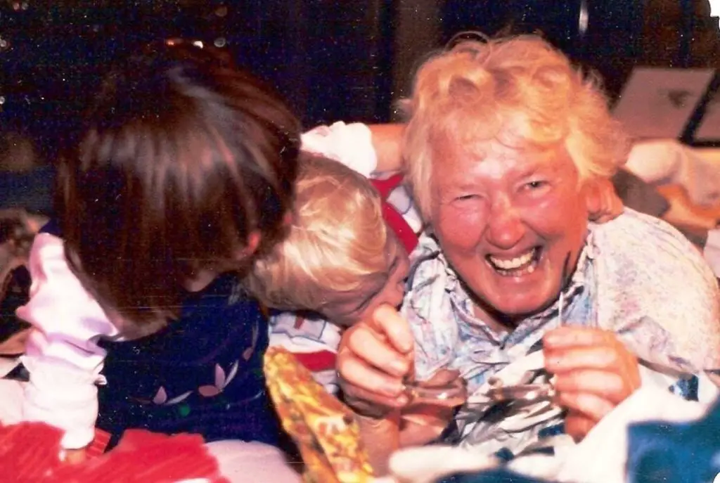 A grandma lays on the floor laughing and playing with two grandchildren amongst wrapping paper