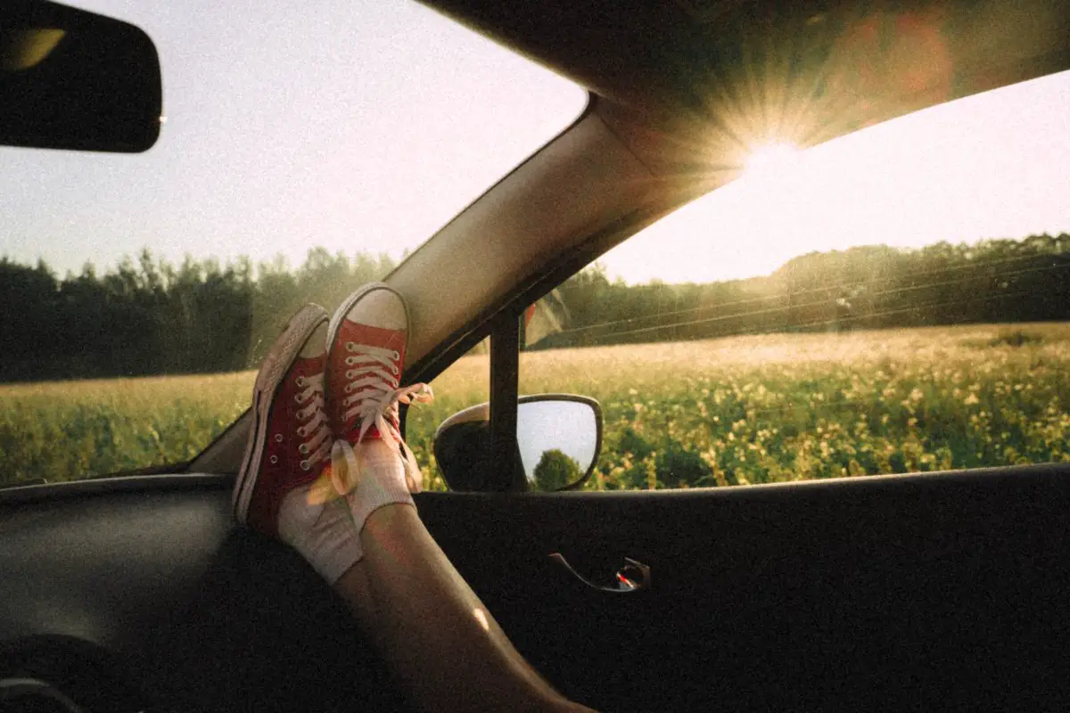 View of the inside of a vehicle. A person rests their feet on the dash with the setting sun shining through the car window in the background