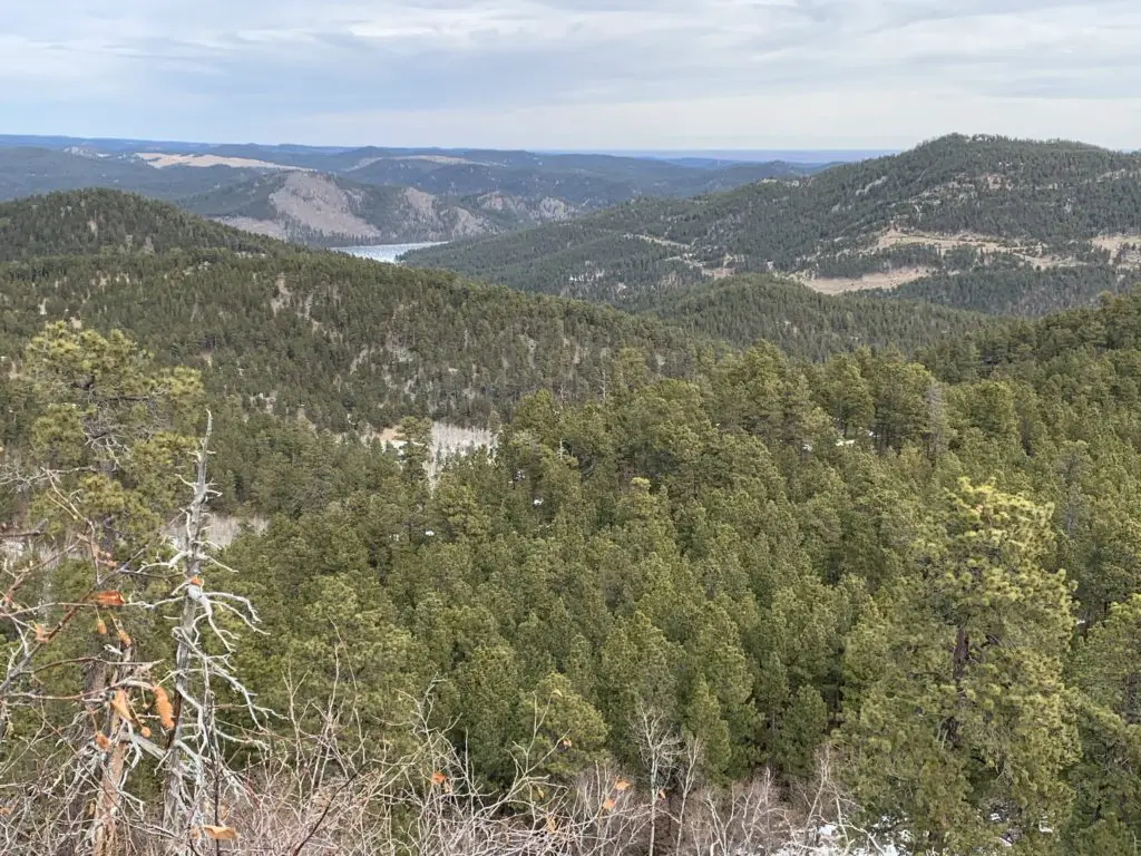 Expansive view over mountains covered in pine trees. A tiny portion of a frozen lake is in the background. 