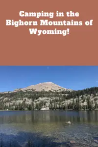 Small lake with rocky, tree-covered mountain peaks in the background. Pin reads, "Camping in the Bighorn Mountains of Wyoming!"