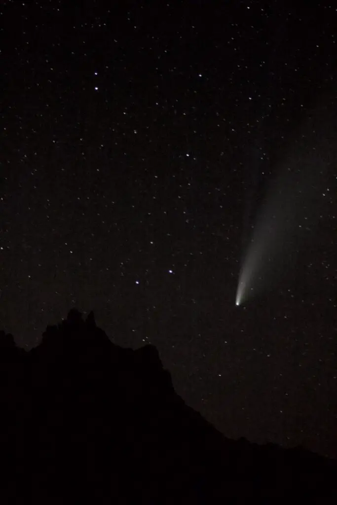 A small, white-colored comet streaks across a dark, night sky dotted with stars