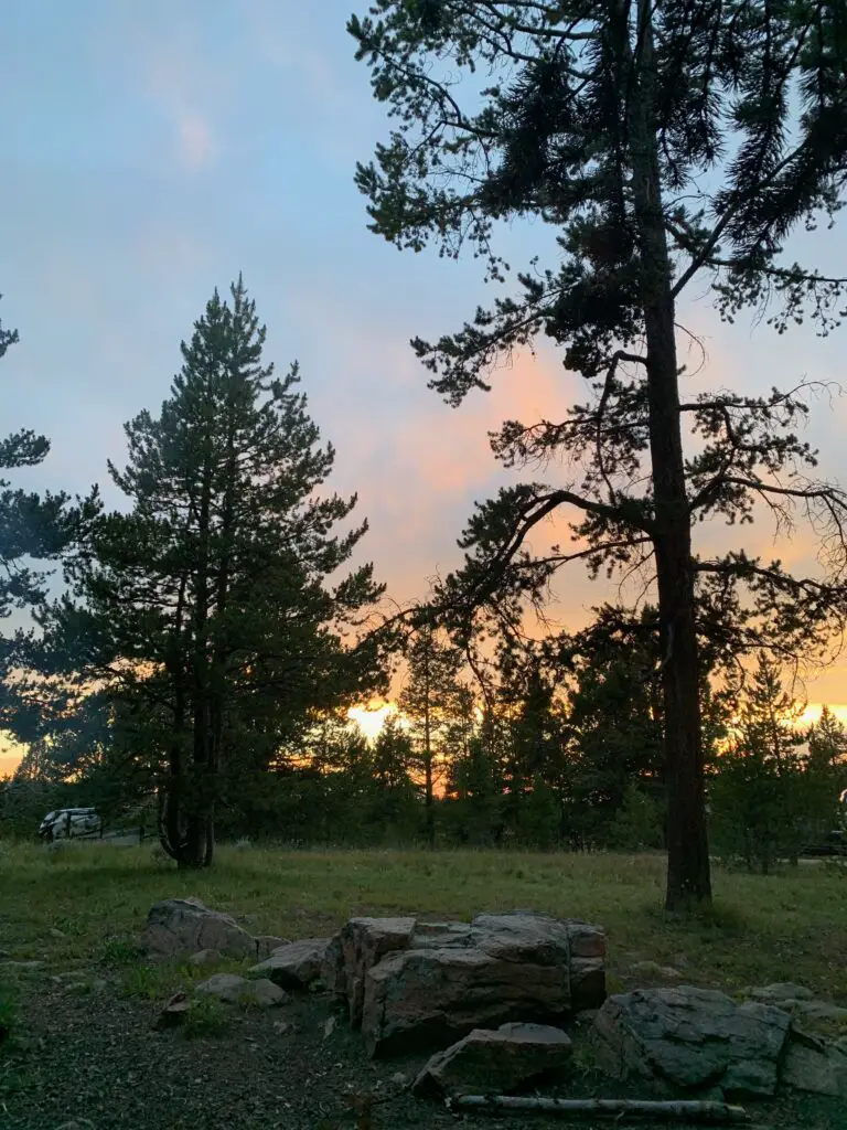A stone fire pit in the foreground with pine trees and a colorful sunset in the background