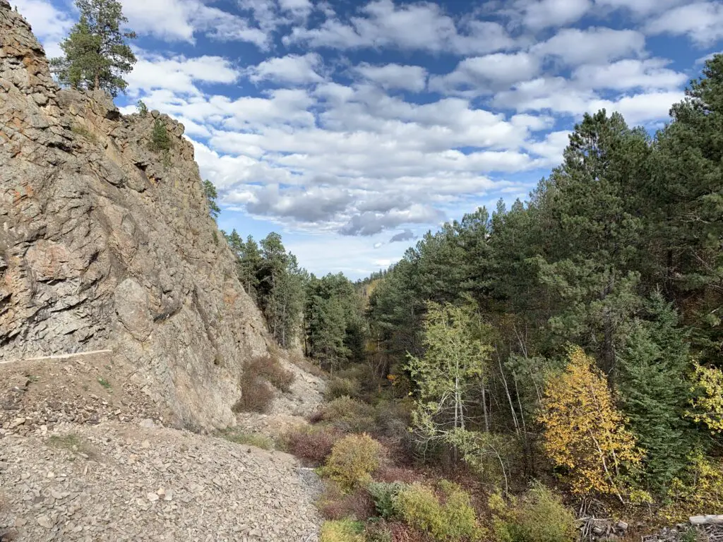 A rocky wall on the left, green and yellow trees on the right, all under a blue sky with puffy, white clouds.